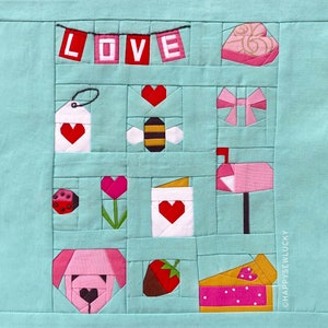 以下が含まれることがあります： 薄い青色の生地に、ハート、蜂、郵便受け、イチゴ、パイ、犬、花、テントウムシ、プレゼント、そして「LOVE」と書かれたバナーなど、バレンタインデーをテーマにした様々なパッチワークのデザインが施されています。