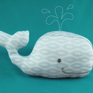 May include: A blue and white whale plush toy with a wave pattern. The whale has a small black eye and a stitched smile. The whale is on a turquoise background with a white dotted line drawing of a water spout above it.