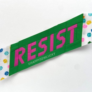 May include: A textile artwork featuring the word "RESIST" in pink capital letters against a green background. The ends of the piece have a white fabric with colourful geometric shapes. The artist's name, "HAPPYSEWLUCKY," is printed at the bottom.