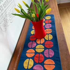 Peut inclure: Un chemin de table matelassé bleu avec un motif géométrique de cercles orange, jaune, rouge et rose. Un bouquet de tulipes jaunes dans un vase rouge est posé sur le chemin de table.