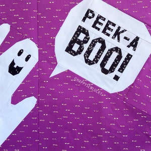 May include: A white fabric ghost with a black star and a smiling face, peeking out from behind a white fabric speech bubble with the text "PEEK-A BOO!" in black. The background is a purple fabric with a repeating pattern of gold eyes.