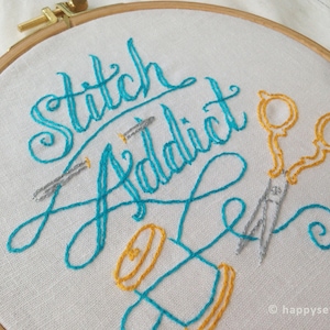 Op de afbeelding: Borduurkunst op een hoepel met de tekst "Stitch Addict" in turquoise draad. De hoepel bevat ook een gouden schaar en een spoel met draad in geel en turquoise.