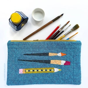 Peut inclure: Une trousse à crayons en denim bleu avec une fermeture à glissière. La trousse a un design matelassé et présente des images brodées de pinceaux et de crayons. Le texte "The depth of your loving is the only way to", "depth of breath", et "was not a day in loving" est brodé sur la trousse.