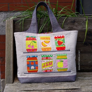 Może przedstawiać: Jasno szara torba typu tote z denimowymi uchwytami prezentuje patchworkowy wzór z kolorowych kwadratów tkaniny przedstawiających słoiki z dżemem. Słoiki mają różne wzory owoców, w tym wiśnie, cytryny, pomarańcze i truskawki. Słowo "JAM" jest nadrukowane na jednym ze słoików.