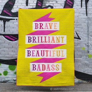 May include: A bright yellow quilt with a pink ribbon design. The quilt features the words "BRAVE," "BRILLIANT," "BEAUTIFUL," and "BADASS" in colourful lettering. The quilt is displayed against a brick wall with graffiti.
