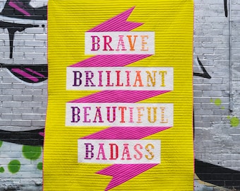 Puede incluir: Un edredón amarillo vibrante con un diseño de cinta rosa que presenta las palabras "BRAVE", "BRILLIANT", "BEAUTIFUL" y "BADASS" en letras coloridas. El edredón se exhibe contra una pared de ladrillos con graffiti.