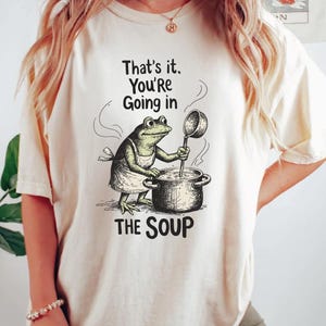 A woman wearing a white t-shirt with a graphic of a frog cooking soup and the text "That's it. You're going in THE SOUP" printed on it.