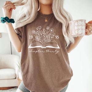 May include: A brown t-shirt with a white floral design emerging from an open book. The text "1995" is above the book, and "chapter thirty" is written below. The shirt is a casual style.