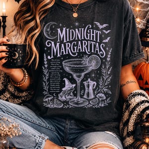 May include: A charcoal gray t-shirt featuring a light purple graphic print with the text Midnight Margaritas. The design includes a margarita glass, a frog wearing a witch hat, a coffee pot, mushrooms, stars, and a crescent moon.