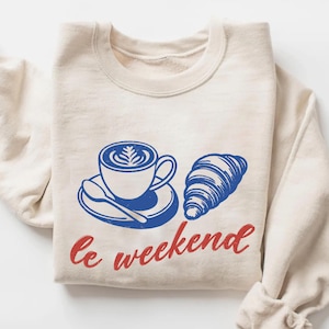 A beige sweater with a graphic design depicting a cup of coffee and a croissant, along with the text "le weekend" printed on it.
