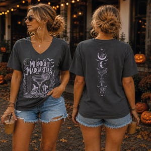 May include: A charcoal gray V-neck t-shirt featuring a white graphic print. The front displays the text Midnight Margaritas with a margarita glass, a witch hat, and a coffee pot. The back shows a vertical design of moons, stars, and icons.
