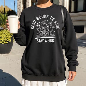 A woman wearing a black sweatshirt with a white graphic design and text, standing on a sidewalk with a white cup in her hand.