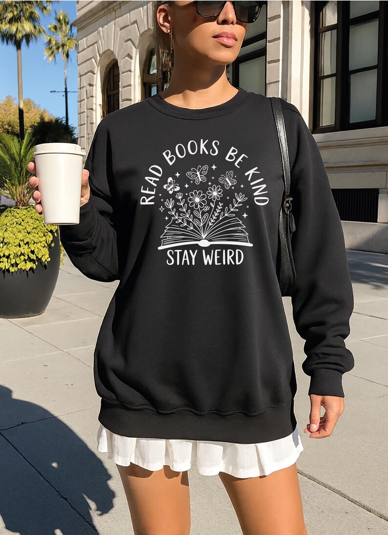 A woman wearing a black sweatshirt with a white graphic design and text, standing on a sidewalk with a white cup in her hand.