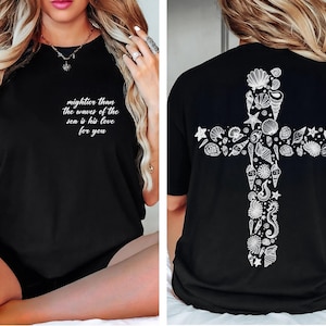 The image is a collage of two photos. In the left photo, a woman with long blonde hair is sitting on a bed, wearing a black t-shirt with a cross design and text on it. In the right photo, the back of the same t-shirt is shown, featuring a cross design with various sea-themed elements such as shells, starfish, and seaweed.