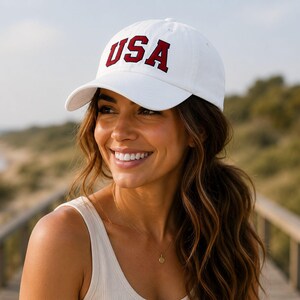 A woman wearing a white baseball cap with the word "USA" on it, smiling and looking off to the side.