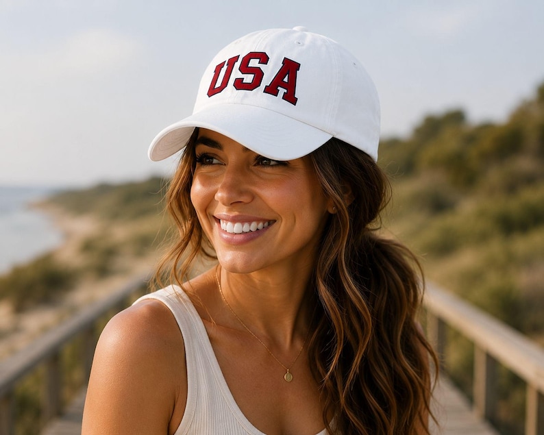 A woman wearing a white baseball cap with the word "USA" on it, smiling and looking off to the side.