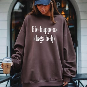 A woman wearing a brown sweatshirt with the text "life happens dogs help" printed on it, holding a drink and standing in front of a building.