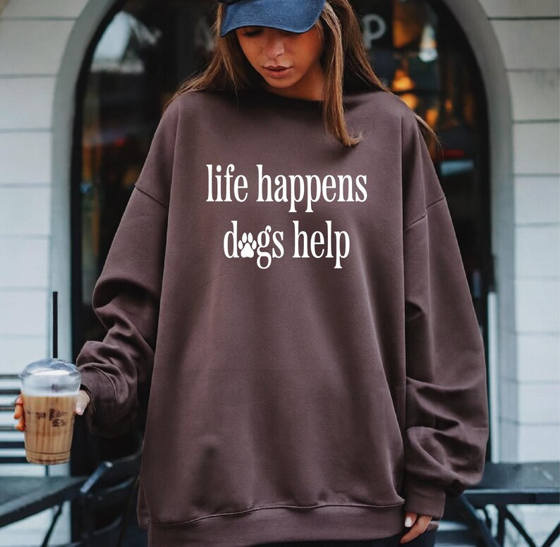 A woman wearing a brown sweatshirt with the text "life happens dogs help" printed on it, holding a drink and standing in front of a building.