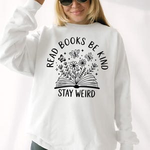 A woman wearing a white sweatshirt with the text "Read Books Be Kind Stay Weird" printed on it, smiling and posing for the camera.