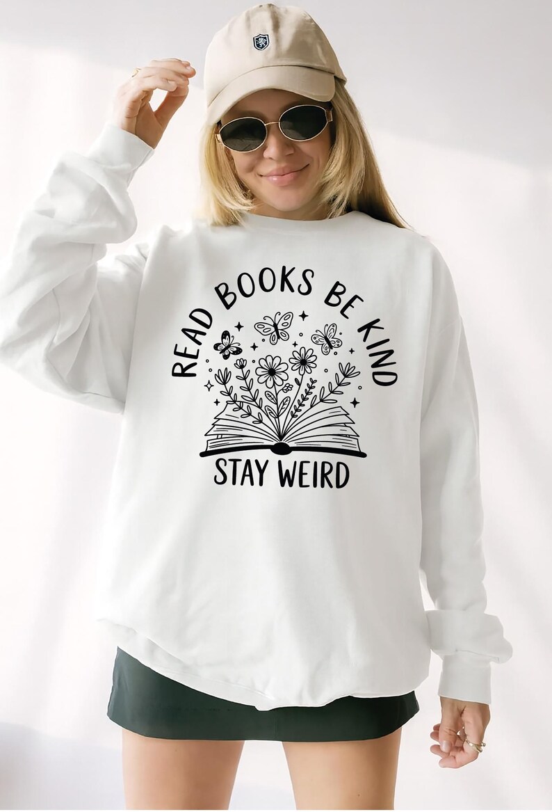 A woman wearing a white sweatshirt with the text "Read Books Be Kind Stay Weird" printed on it, smiling and posing for the camera.
