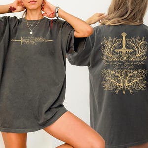 May include: Dark grey t-shirt with a gold sword and floral design on the front and back. The back of the shirt features the text "You do not fear. You do not falter. You do not yield."