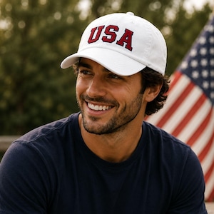 A man wearing a white baseball cap with the word "USA" on it, smiling and looking to the side.