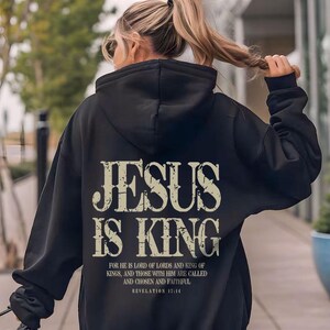 A woman wearing a black hoodie with the text "JESUS IS KING" printed on it, walking on a sidewalk.