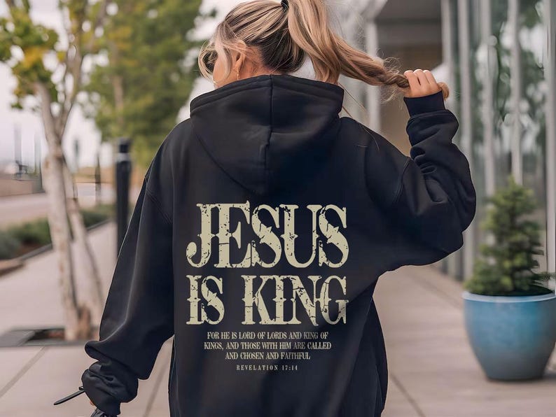 A woman wearing a black hoodie with the text "JESUS IS KING" printed on it, walking on a sidewalk.