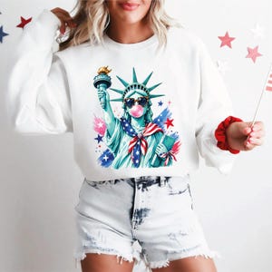 May include: White sweatshirt featuring a graphic of the Statue of Liberty wearing sunglasses and blowing a bubblegum bubble. The Statue is in blue and green with red, white, and blue accents. The design includes stars and a torch.
