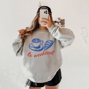 A person is taking a selfie with a smartphone, wearing a grey sweatshirt with a graphic design and the text "Le weekend" printed on it.