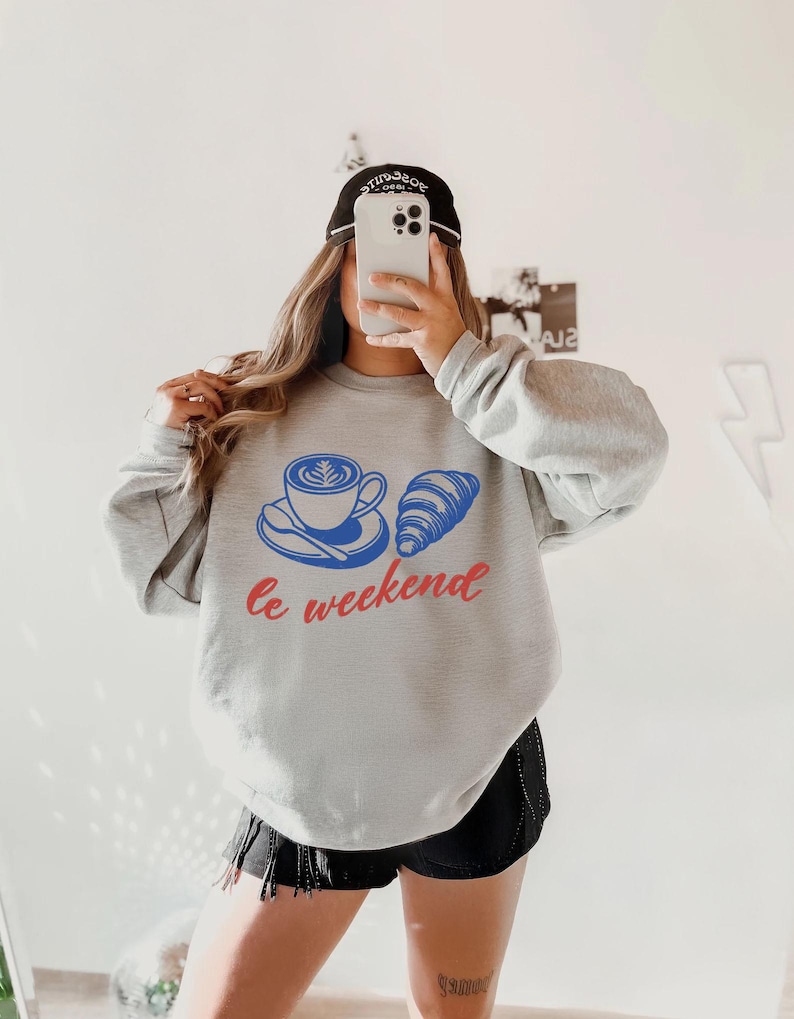 A person is taking a selfie with a smartphone, wearing a grey sweatshirt with a graphic design and the text "Le weekend" printed on it.