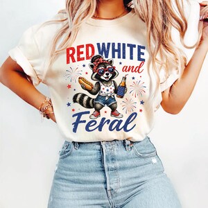 May include: A cream-colored t-shirt featuring a graphic of a raccoon wearing heart-shaped sunglasses and a bandana, holding a hot dog and a bottle. The text reads RED WHITE and Feral, surrounded by fireworks and stars in red, white, and blue colors.