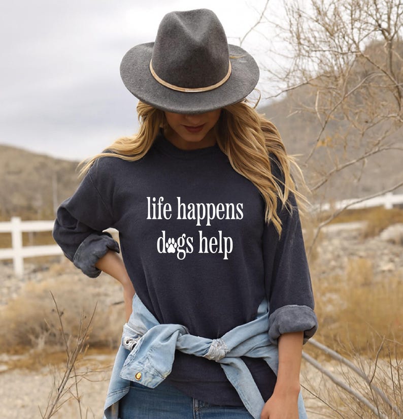 A woman wearing a black hat and a black sweatshirt with the text "life happens dogs help" stands in a field with trees in the background.
