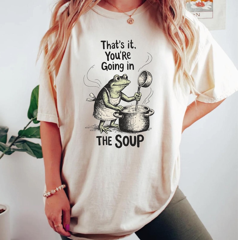 A woman wearing a white t-shirt with a graphic of a frog cooking soup and the text "That's it. You're going in THE SOUP" printed on it.