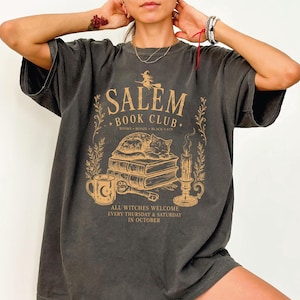 A person wearing a gray t-shirt with a graphic design featuring a cat, books, and a witch on a broomstick.