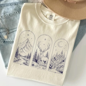 May include: A cream-colored t-shirt featuring three vertical arched panels with dark blue line art illustrations. The left panel shows a mountain landscape, the center shows a deer in a forest, and the right shows a castle under a crescent moon.