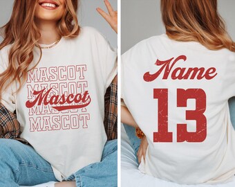 May include: Cream-coloured t-shirt featuring the word "MASCOT" in a stacked design, with a red script "Mascot" in the centre. The back of the shirt displays the word "Name" in red script above the number "13" in a bold, red font. The shirt has a casual, oversized fit.