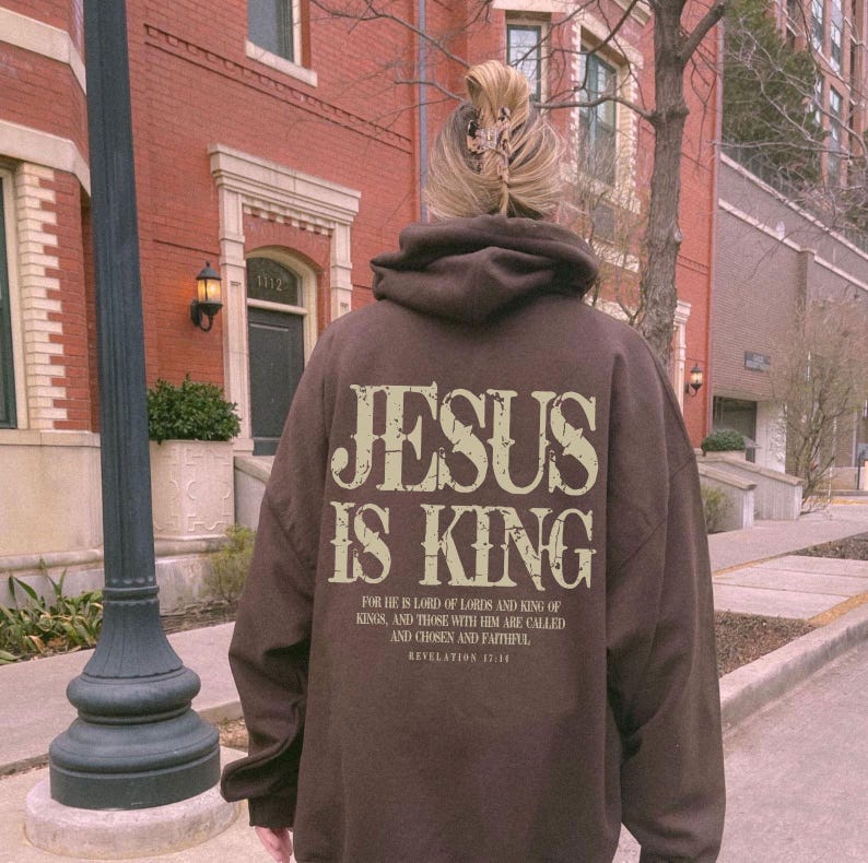 A person wearing a brown hoodie with the text "JESUS IS KING" printed on it, standing in front of a brick building.