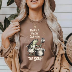 A woman wearing a brown t-shirt with a cartoon frog holding a ladle and standing in a pot, with the text "That's it. You're going in the soup" printed on it.