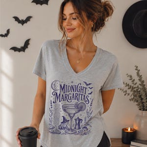 May include: A light gray V-neck t-shirt featuring a dark blue graphic print with the text "MIDNIGHT MARGARITAS," a margarita glass, a frog wearing a witch hat, a moka pot, mushrooms, stars, and a crescent moon, all arranged in a detailed design.