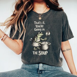 A woman wearing a gray t-shirt with a graphic of a frog cooking in a pot and the text "That's it, You're going in the soup" printed on it.