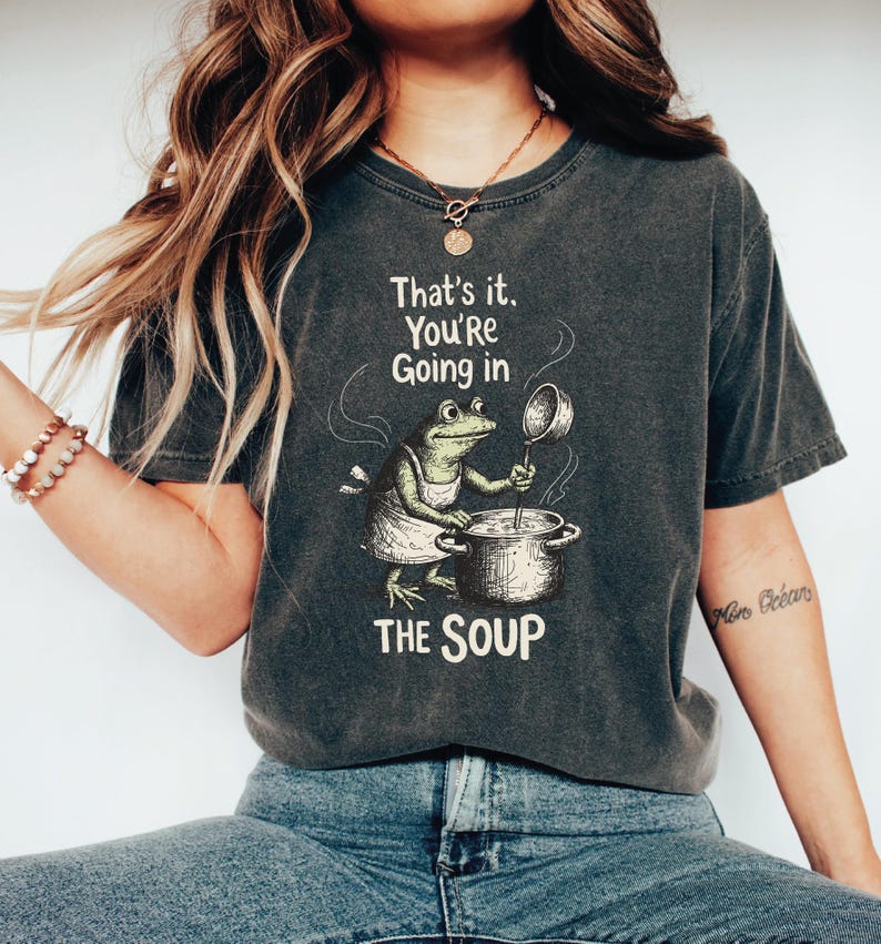 A woman wearing a gray t-shirt with a graphic of a frog cooking in a pot and the text "That's it, You're going in the soup" printed on it.