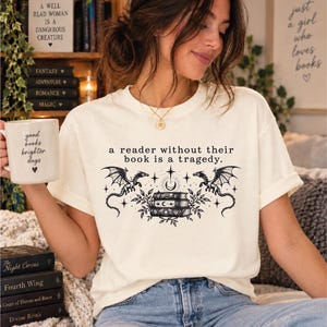 May include: A cream-colored t-shirt featuring a black graphic of three stacked books, two dragons, stars, and a crescent moon. The text reads, "a reader without their book is a tragedy." A white mug with the text "good books brighter days" is held nearby.