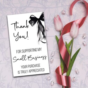 a white card with a black ribbon and pink tulips, along with a red ribbon and small white flowers, all arranged on a light pink background.