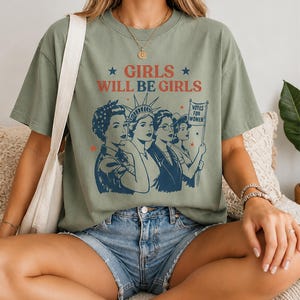 A woman wearing a green t-shirt with a graphic design of four women and the text "GIRLS WILL BE GIRLS" printed on it.