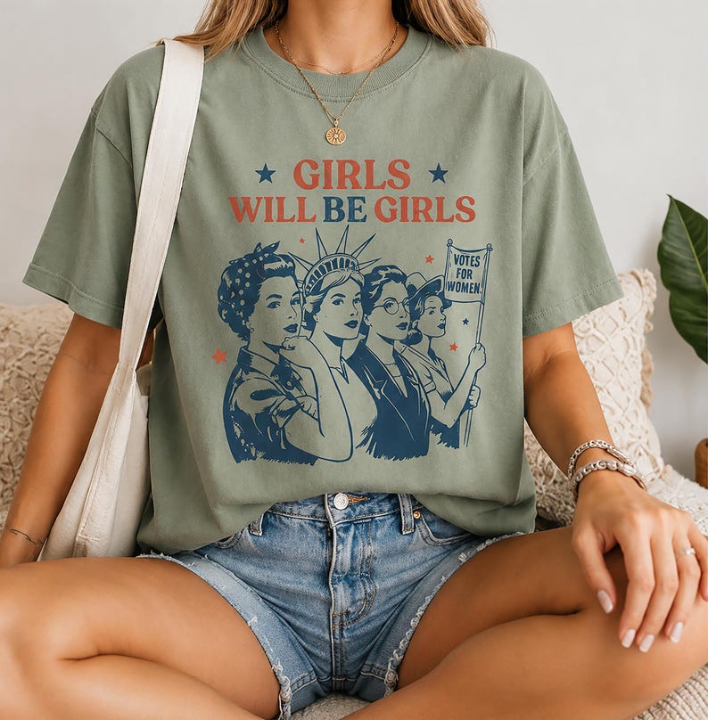 A woman wearing a green t-shirt with a graphic design of four women and the text "GIRLS WILL BE GIRLS" printed on it.
