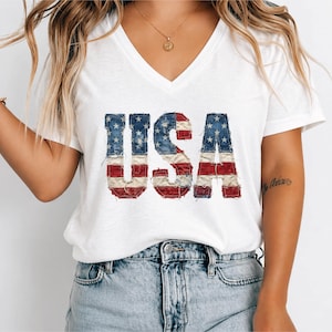 May include: White V-neck t-shirt with the word "USA" in a distressed, patriotic design. The letters are made to look like the American flag, with red and white stripes and blue stars.