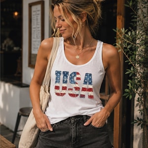 May include: White tank top with the word "USA" in a distressed American flag design. The top is paired with black denim shorts. A beige tote bag is slung over the shoulder.