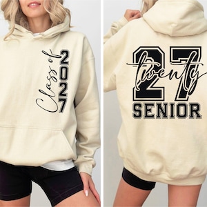 May include: A cream-colored hooded sweatshirt featuring black text. The front displays vertical text reading Class of 2027. The back features a large number 27 with the word twenty written in script across it and the word SENIOR printed below in block letters.