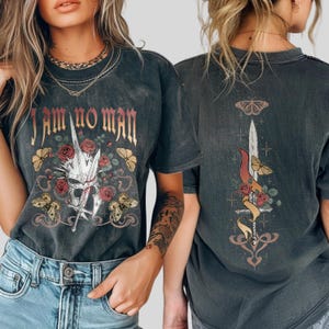 May include: Dark gray t-shirt with the text "I AM NO MAN" in gold lettering. The front graphic features a skull with a sword, roses, and butterflies. The back of the shirt has a sword, roses, butterflies, and a ribbon design.